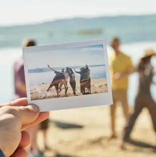 A hand holding a Polaroid photo of friends on a beach, capturing a joyful vacation memory with Sunset Bliss Travels.
