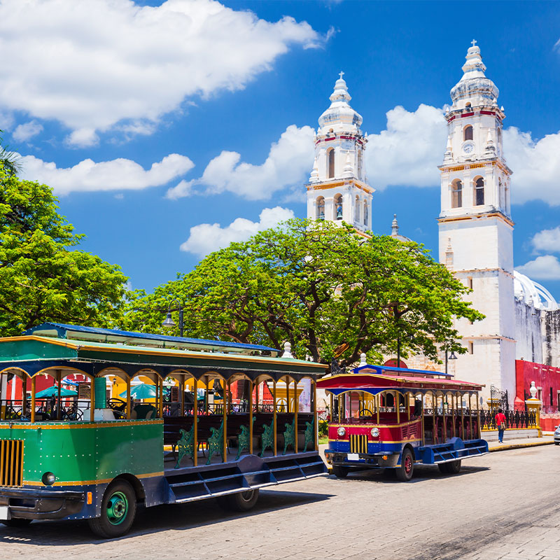 Colorful tourist trolleys parked in front of the historic, white-towered Campeche Cathedral under a bright blue sky in Mexico.