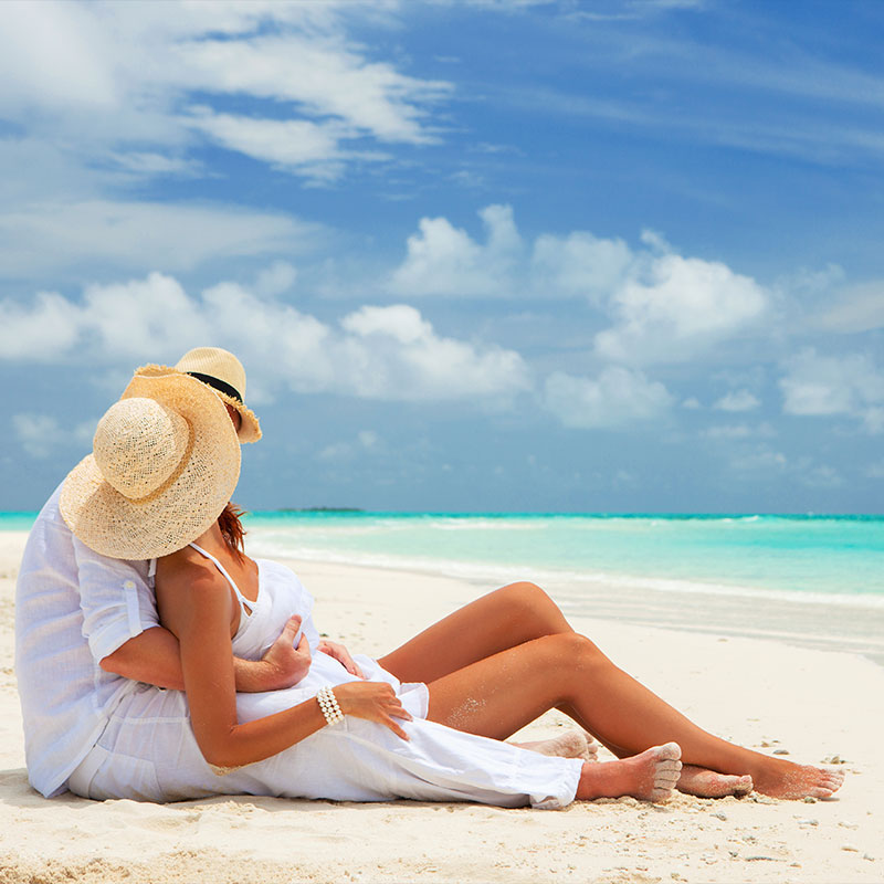 A couple in white attire sitting on a serene white sand beach, looking out at the bright Carribean ocean.
